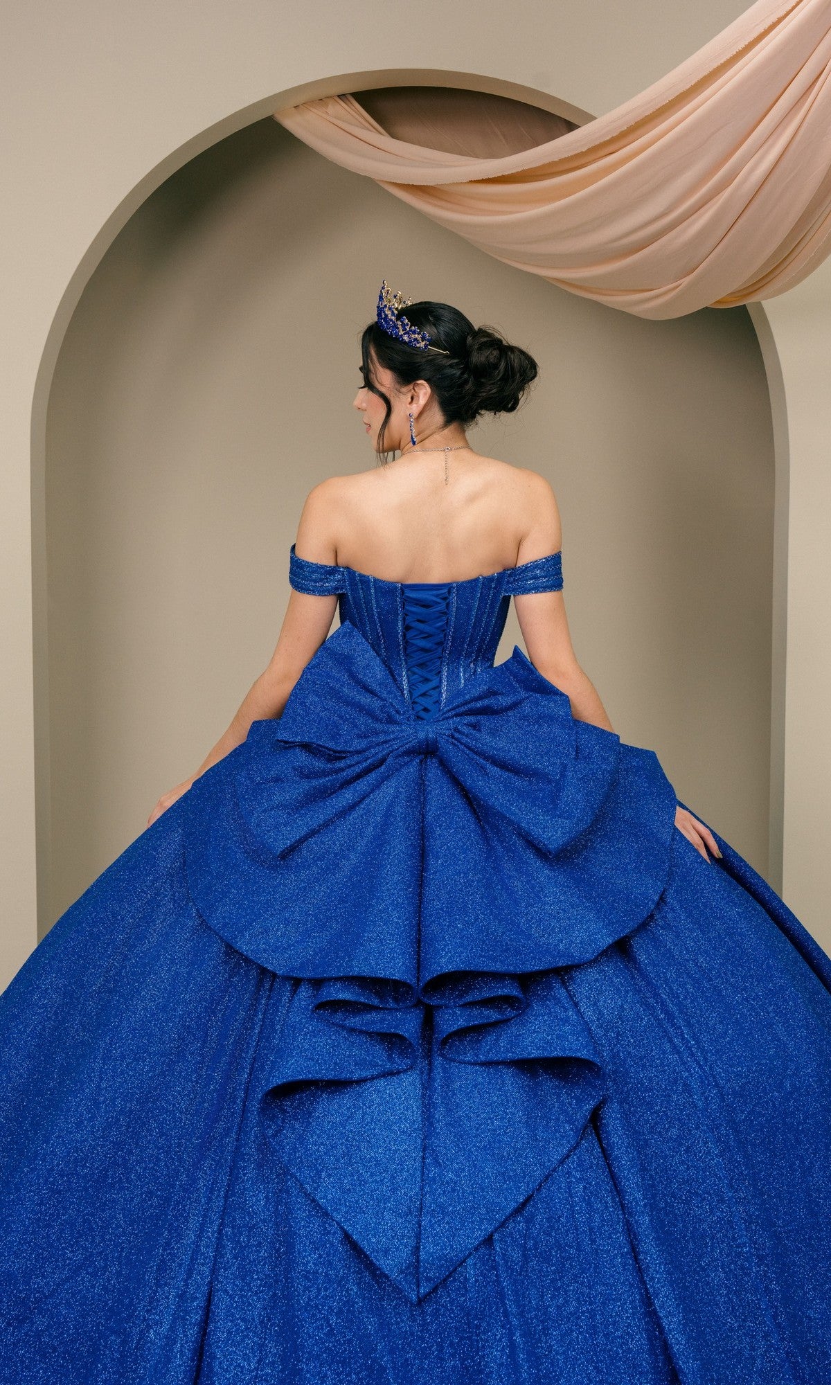 A woman in the Quinceanera Dress 8460JQ by Cinderella Couture, wearing a tiara, stands with her back to the camera, framed by an arched backdrop and draped fabric.
